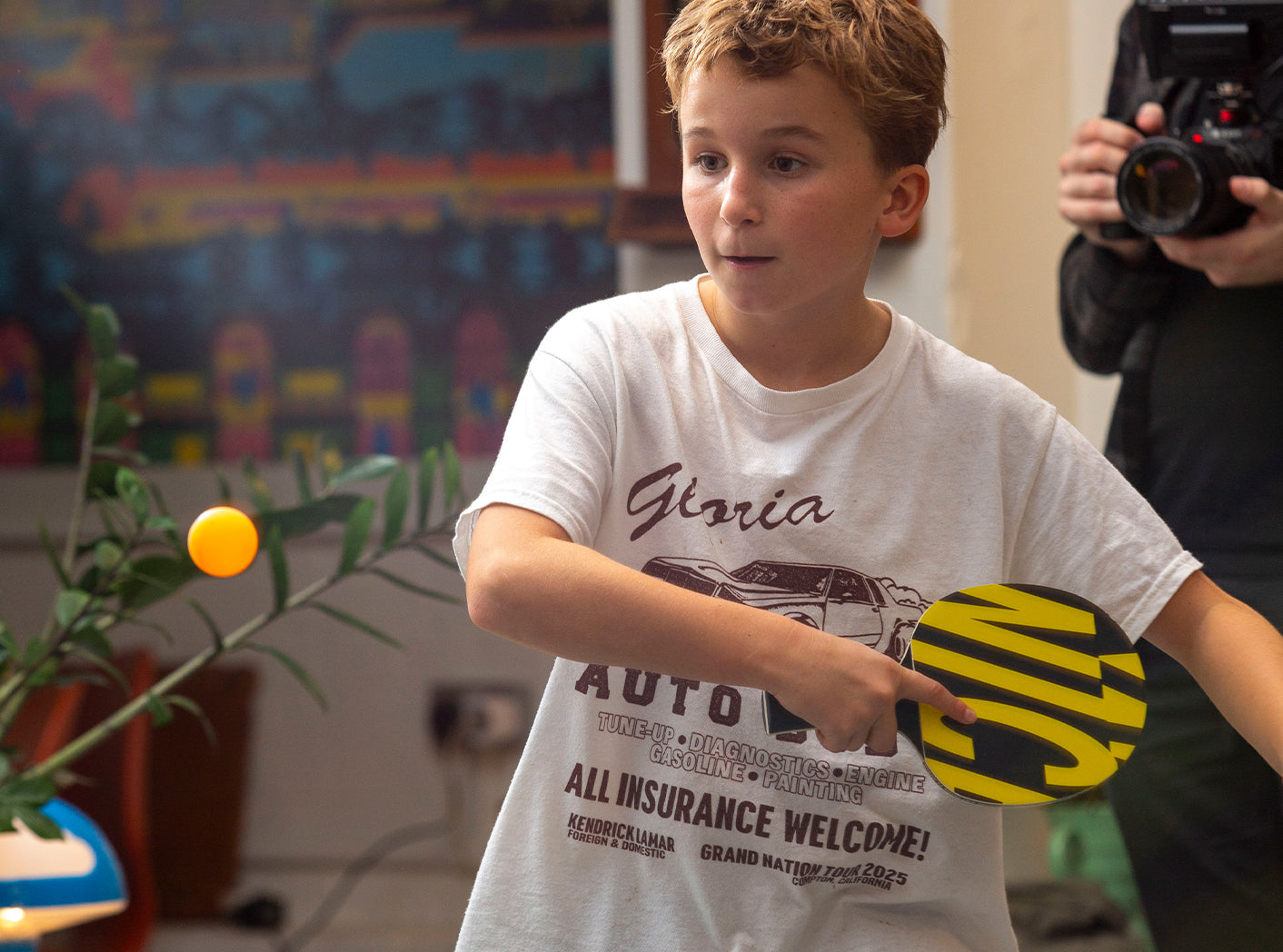 Boy holding a ping pong paddle with a camera in the background, in an indoor setting.